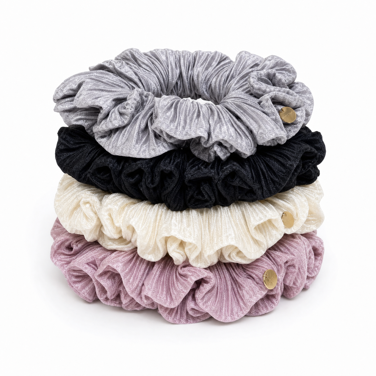 Vertical stack of four ruffled silky satin scrunchies in dove grey, black, cream, and plum featuring gold button accents.